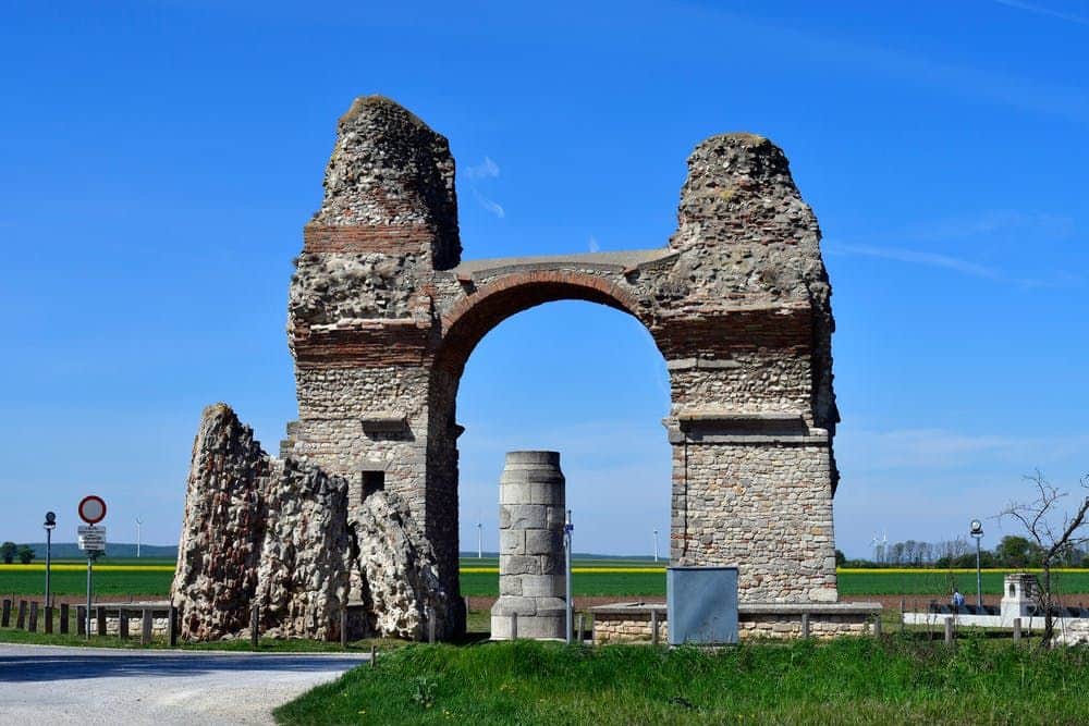 Anciennes ruines d'arches en pierre avec une grande arche centrale et des colonnes qui l'entourent, sur fond de ciel bleu clair et de paysage verdoyant.