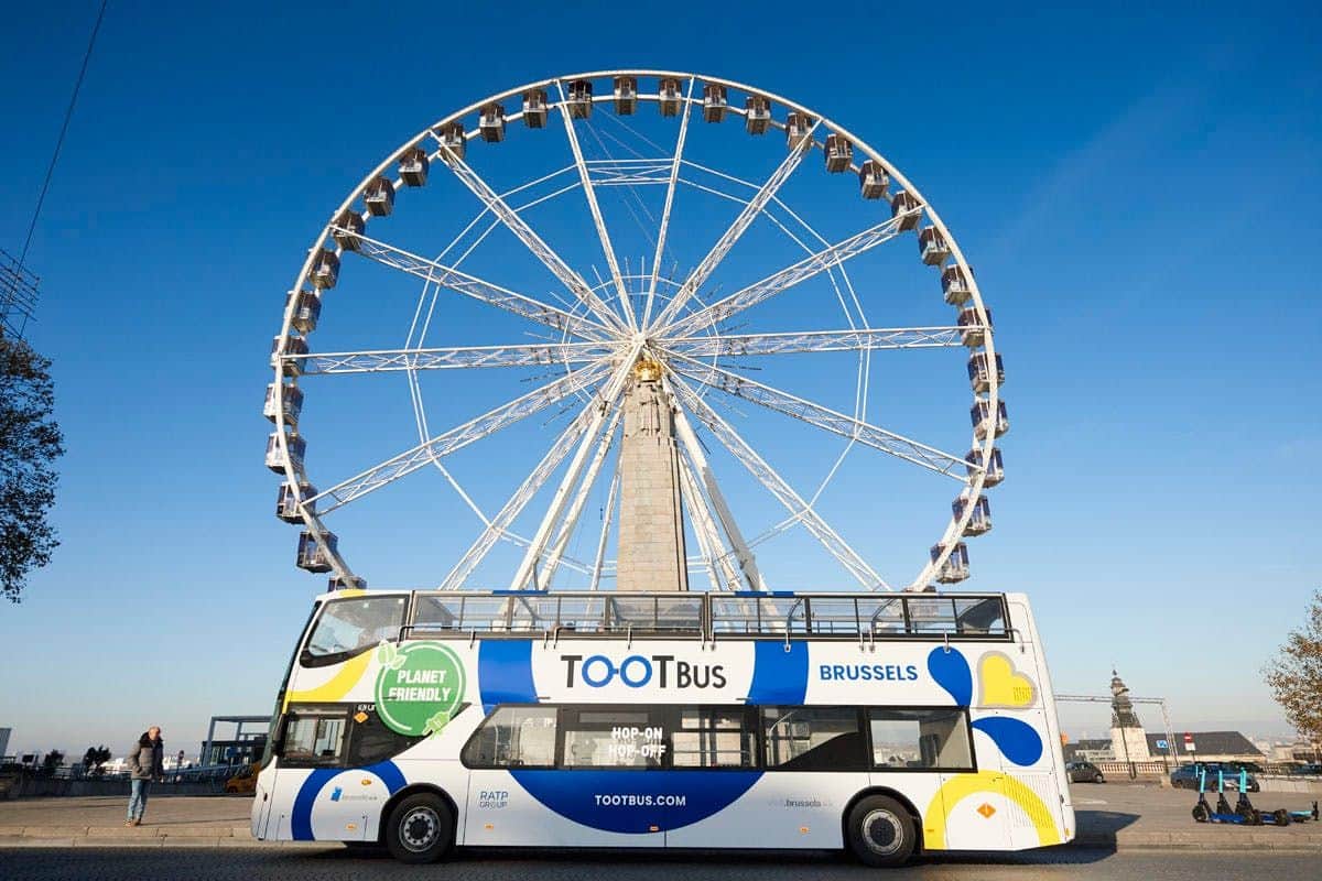 Un bus touristique à impériale portant l'inscription "TOOTBus Brussels" stationne devant une grande roue dans un ciel bleu limpide.
