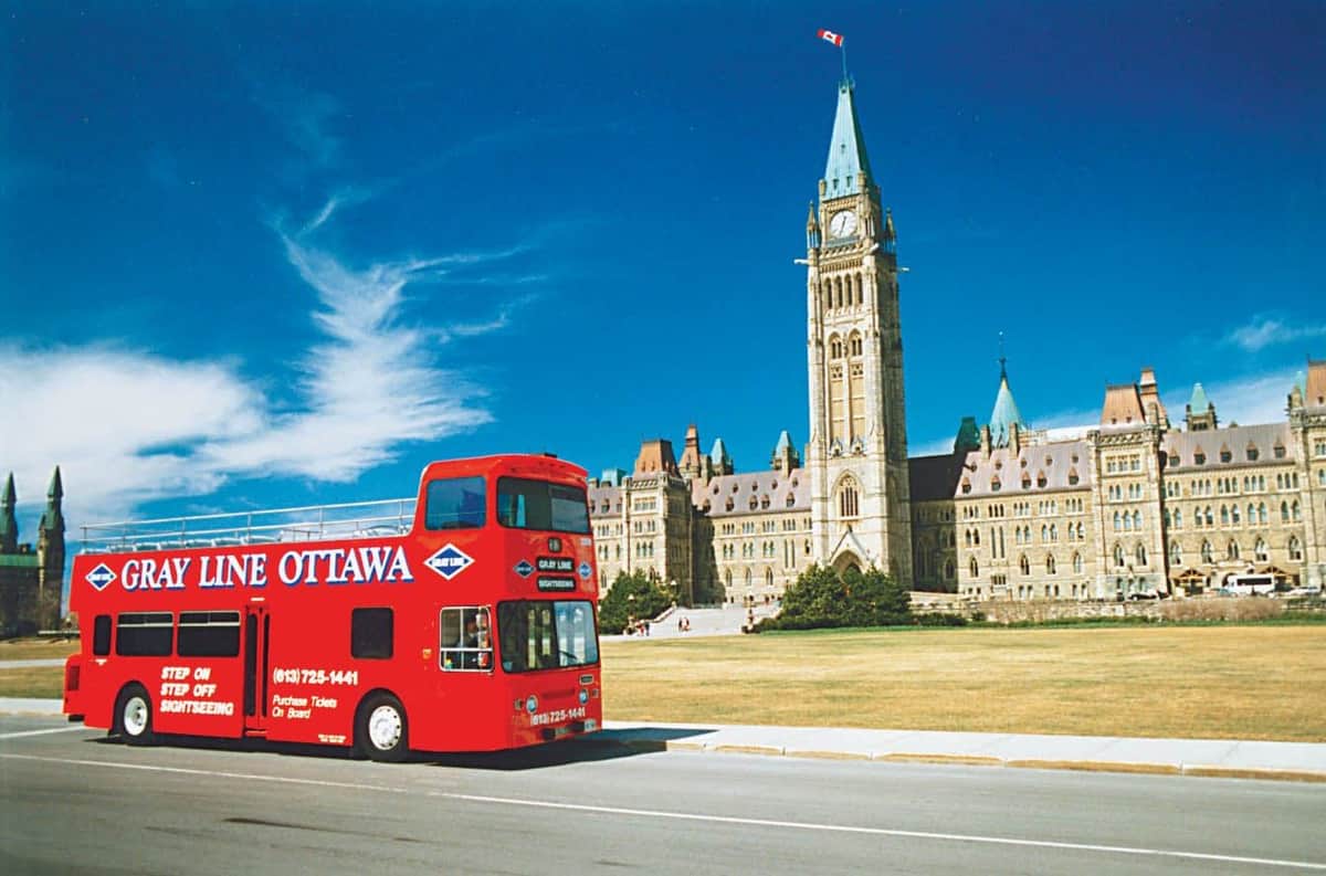 Visite d'Ottawa : Bus hop-on hop-off Un bus touristique rouge à impériale portant l'inscription "OTTAWA" passe devant un grand bâtiment historique doté d'une haute tour d'horloge, sous un ciel bleu clair.