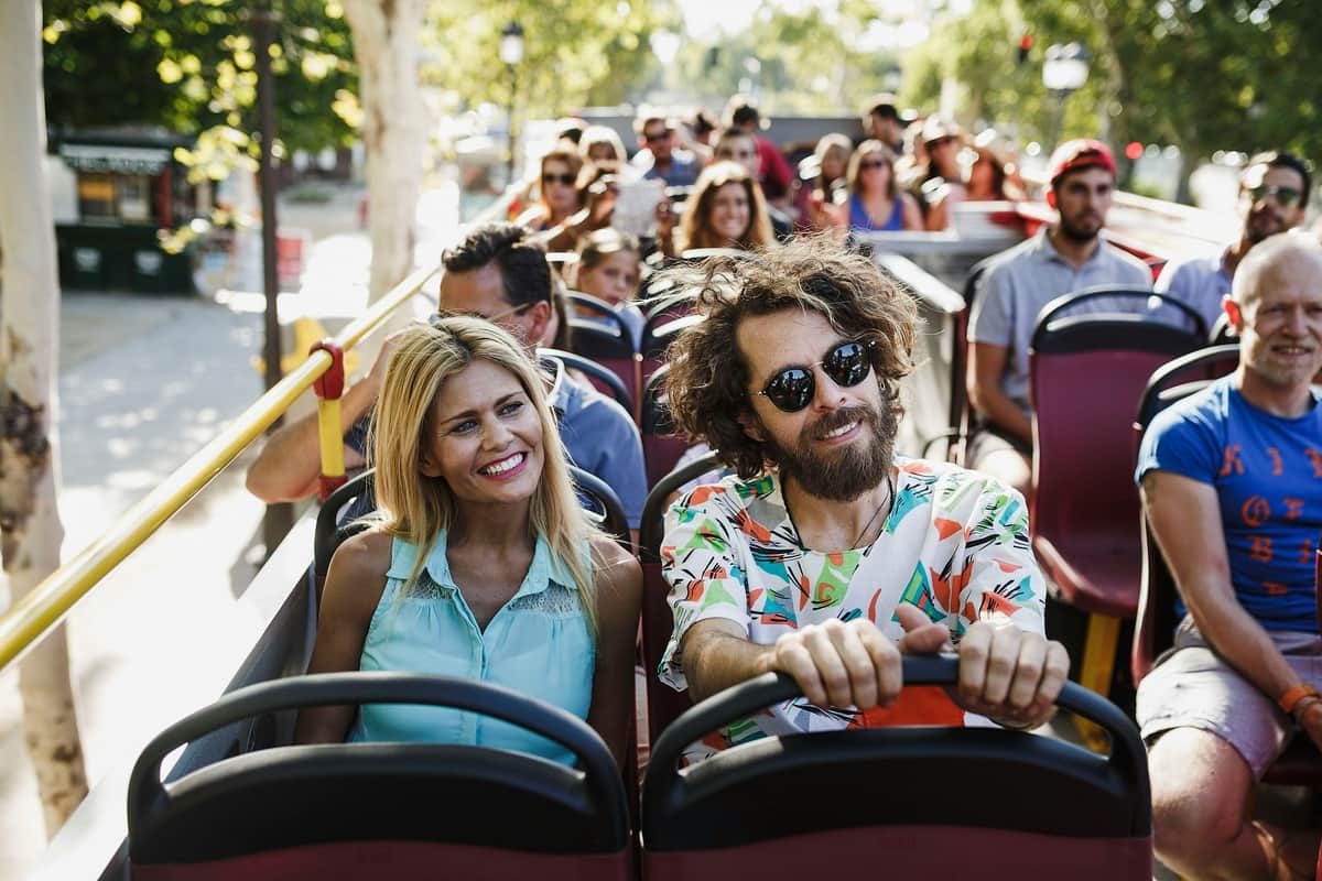 Des personnes assises dans un bus touristique à impériale, souriant et profitant de la promenade par une journée ensoleillée. Des arbres et des lampadaires sont visibles.