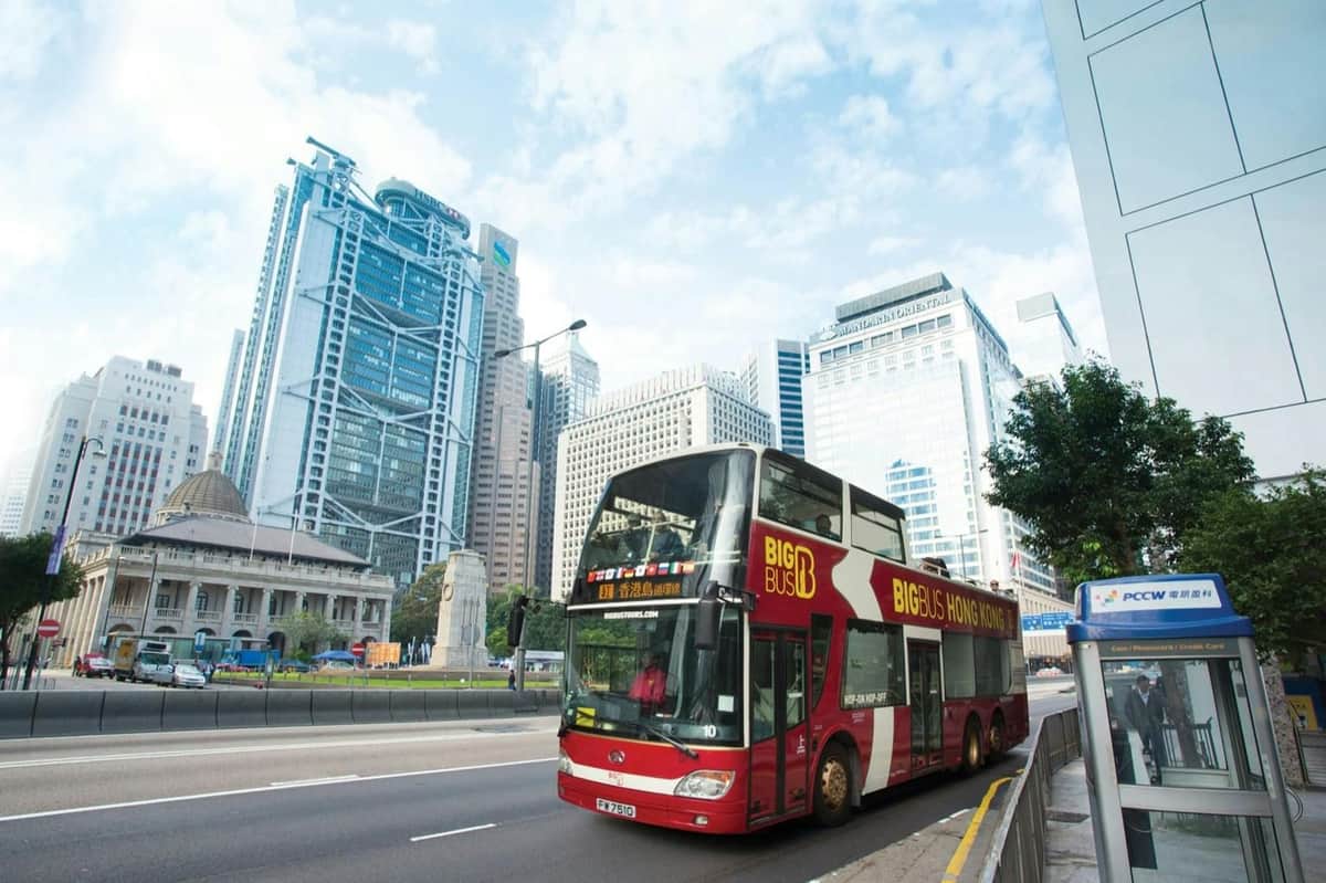 Bus touristique rouge à impériale portant l'inscription "Big Bus Hong Kong" dans une rue de la ville avec des gratte-ciel modernes et un ciel dégagé en arrière-plan.