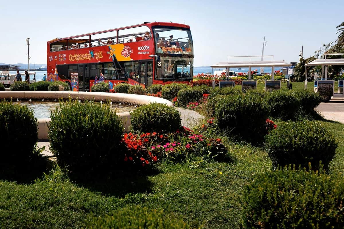 Split : 24 ou 48 heures de Bus Hop-on Hop-off + visite guidée à pied Un bus touristique rouge à toit ouvert, entouré de fleurs colorées et de verdure, avec la mer en arrière-plan.