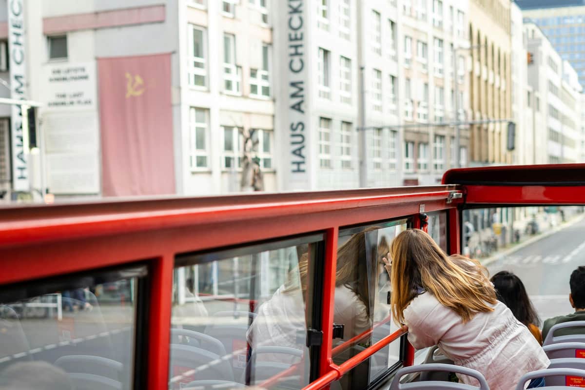 Une femme prend une photo depuis un bus rouge à toit ouvert dans une zone urbaine avec un panneau bien visible "HAUS AM CHECKPOINT CHARLIE".