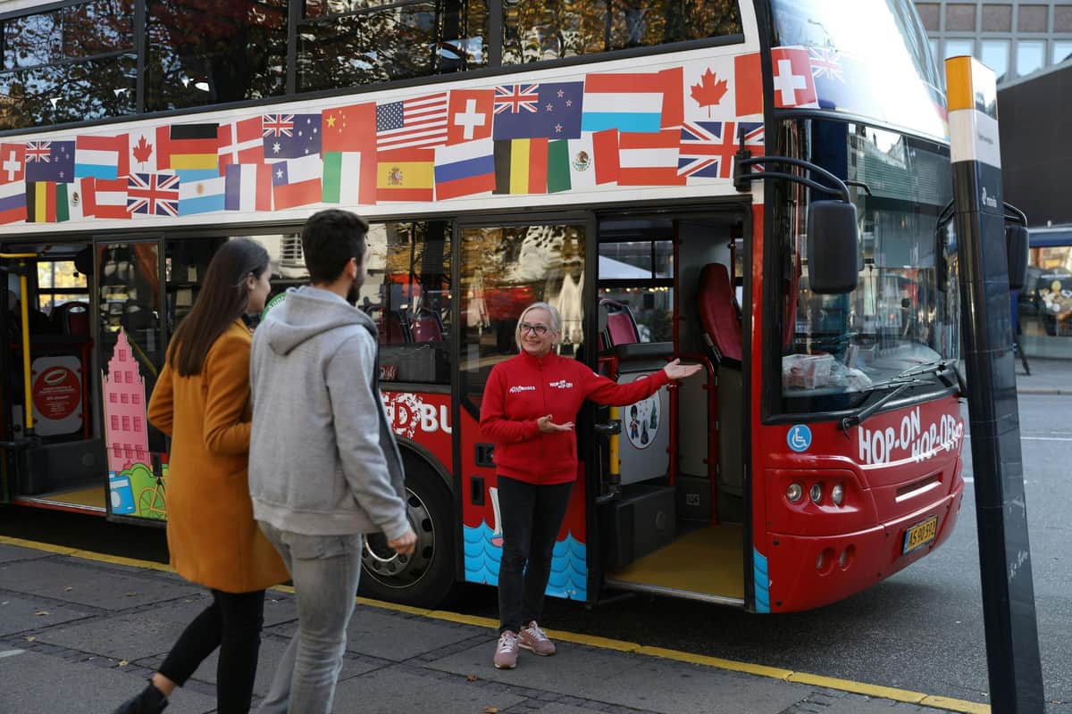 Deux personnes s'approchent d'un guide près de l'entrée d'un bus touristique à impériale décoré de divers drapeaux internationaux.