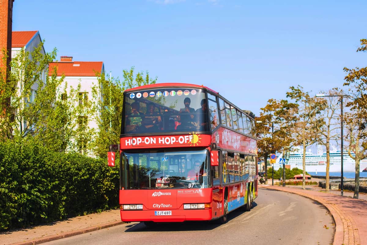 Un bus rouge à impériale "Hop-on Hop-off" circule dans une rue de la ville par une journée ensoleillée. Les passagers sont visibles sur le pont supérieur.