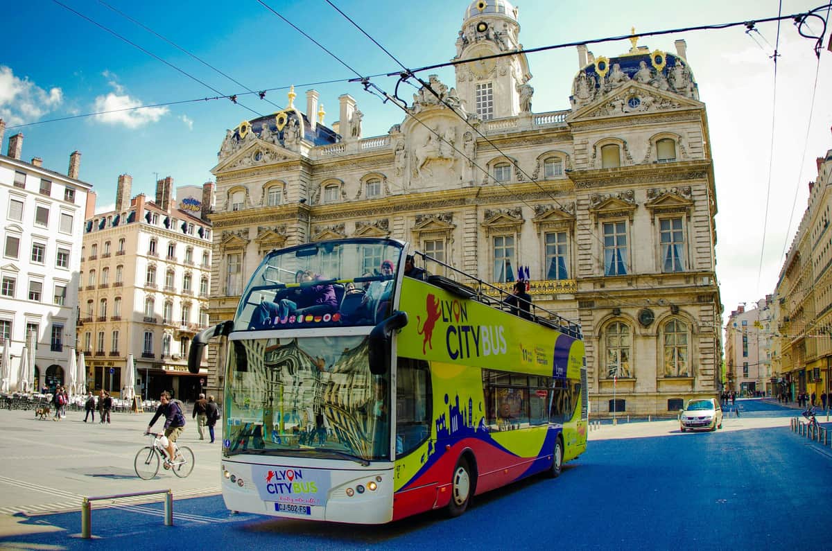 Lyon : Bus Hop-on Hop-off Un bus de la ville de Lyon aux couleurs vives devant un bâtiment historique à l'architecture ornée, par temps clair.