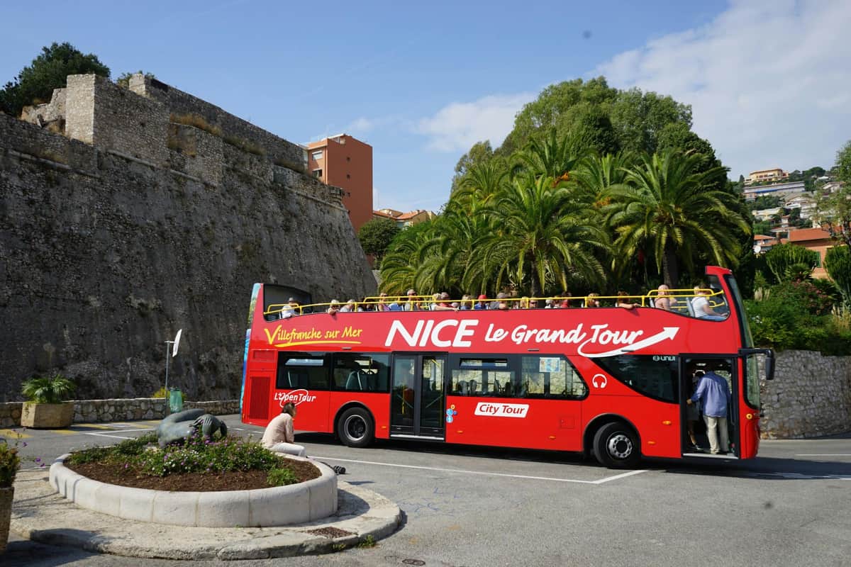 Un bus touristique rouge à impériale portant l'inscription "Nice Le Grand Tour" est garé devant un mur de pierre et des palmiers luxuriants, sous un ciel dégagé.