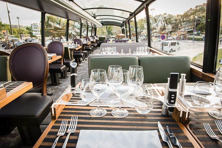 Déjeuner ou dîner gastronomique sur le Bustronome An elegantly set dining table with empty wine glasses and cutlery inside a glass-roofed restaurant bus with exterior city views.