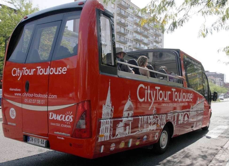 Autobus CityTour rouge avec passagers, toit ouvert, et monuments toulousains représentés sur le côté, circulant près d'un immeuble résidentiel.