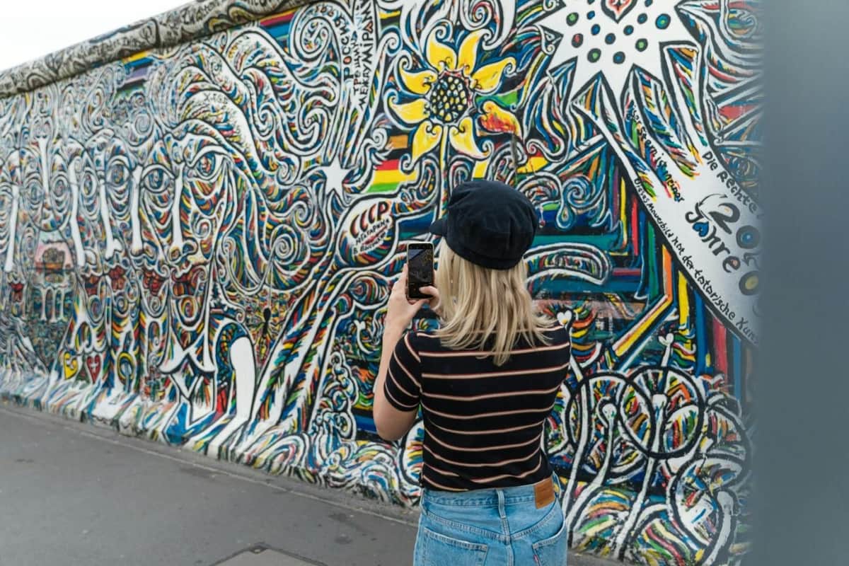 A woman in a striped shirt and black hat takes a photo of a colorful, intricate mural on an outdoor wall.