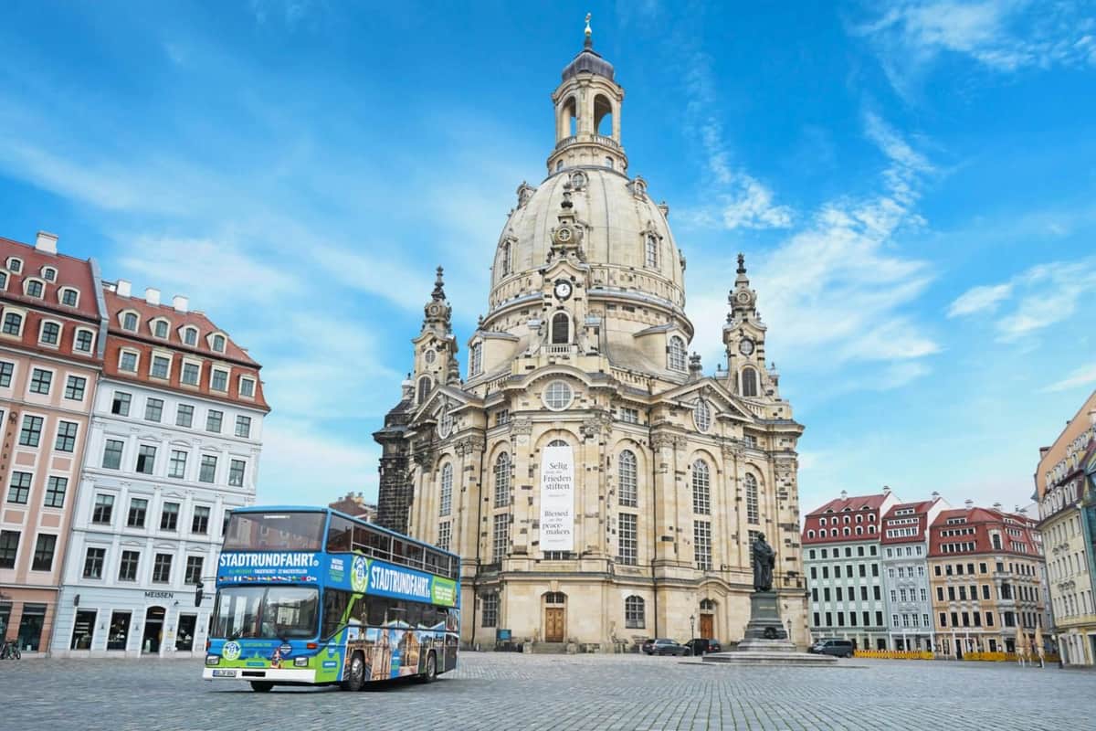 Bus hop-on hop-off Dresde Bus touristique à côté de l'église baroque Frauenkirche de Dresde sous un ciel bleu, avec les bâtiments historiques environnants en arrière-plan.