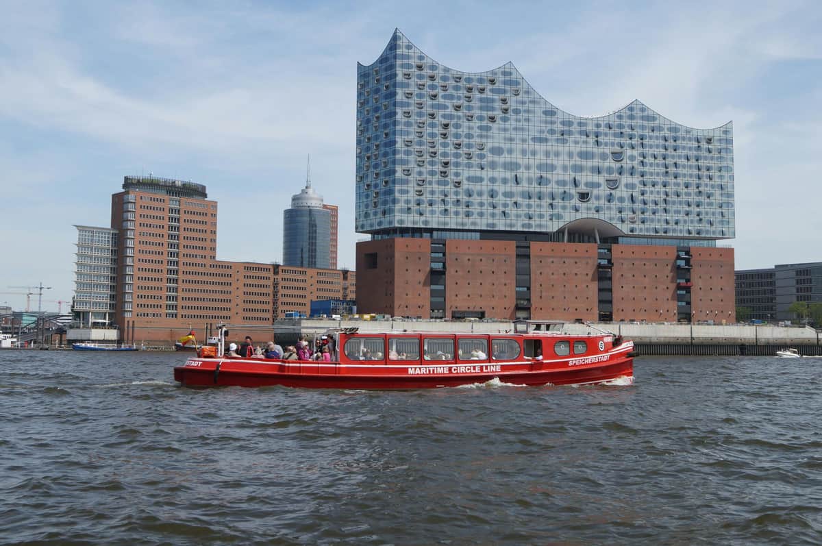 Un bateau rouge nommé "Maritime Circle Line" avec des passagers devant des bâtiments modernes et en brique au bord de l'eau.