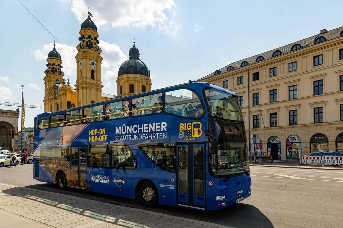 Un bus bleu à impériale stationne devant des bâtiments historiques avec des tours sous un ciel ensoleillé.