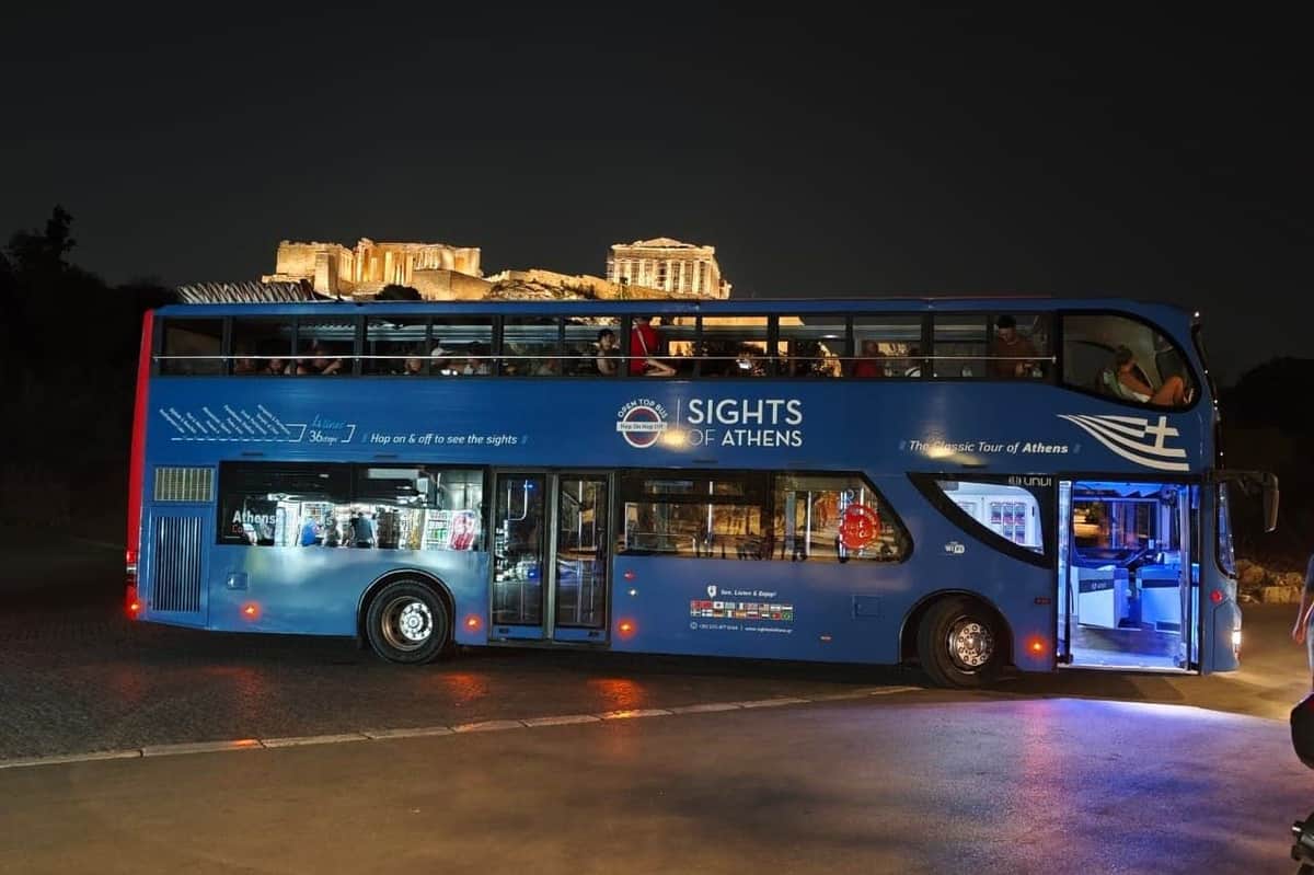 Athènes : Visite guidée en bus de nuit Un bus bleu à impériale intitulé "Sights of Athens" est garé de nuit avec des ruines antiques illuminées visibles à l'arrière-plan.