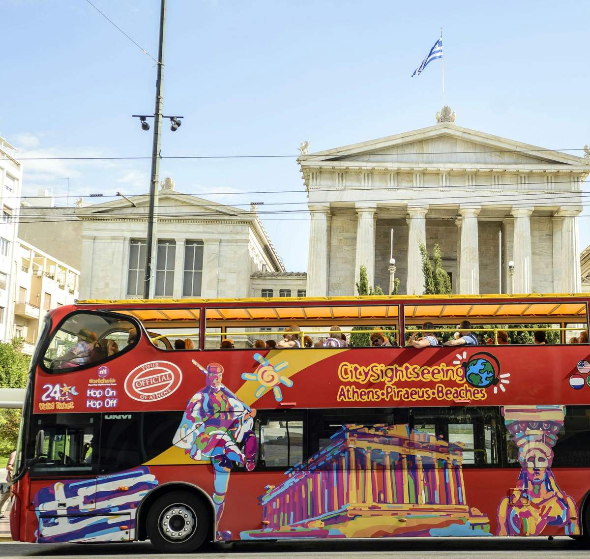 Un bus touristique à impériale, très vivant, sur lequel sont peints les monuments d'Athènes, passe devant l'architecture grecque classique.
