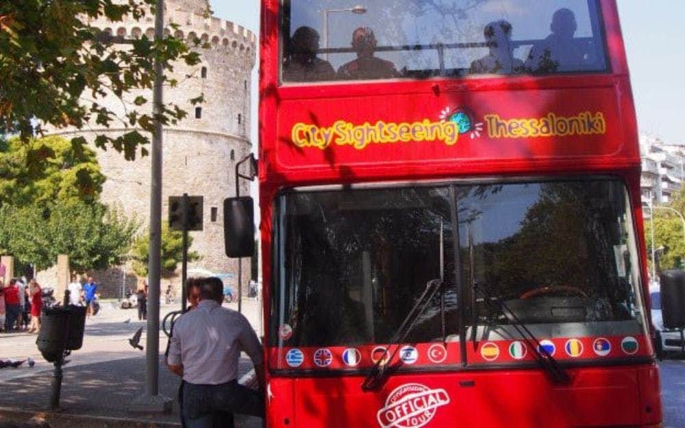 Autobus rouge à impériale près d'une tour historique en pierre. Des personnes sont visibles sur le pont supérieur du bus, des ombres d'arbres se superposent au bus.