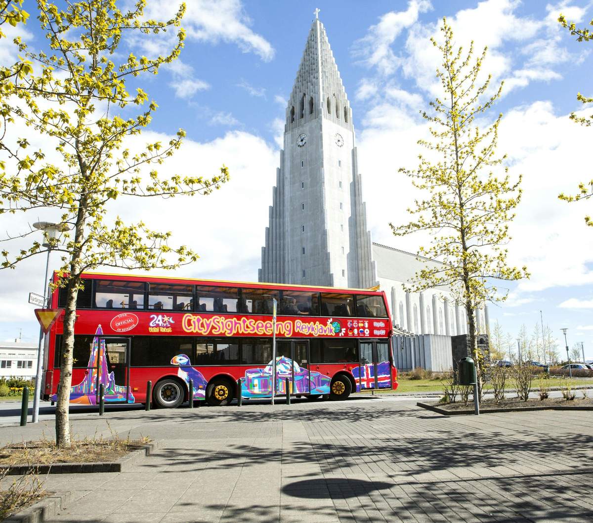 Un bus touristique rouge City Sightseeing devant une église haute et pointue, avec des arbres et un ciel bleu en arrière-plan.