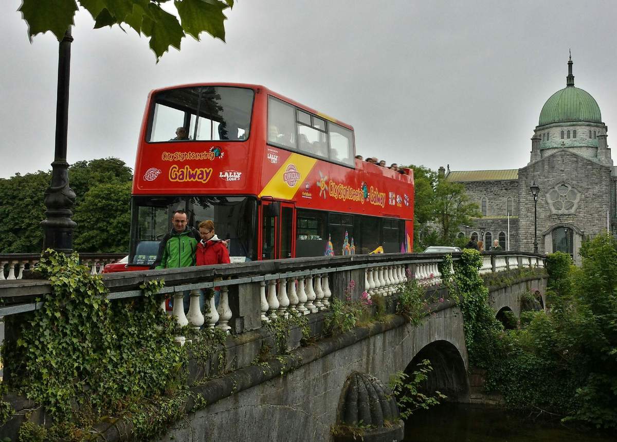 Un bus rouge à impériale passe sur un pont en pierre recouvert de lierre, deux personnes se tenant à la rambarde. Un grand bâtiment en pierre est visible derrière.