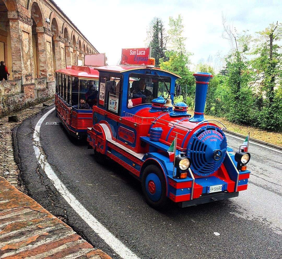 Un train touristique bleu et rouge portant l'inscription "San Luca Express" circule sur une route incurvée près d'une vieille arche en briques, entourée de verdure.