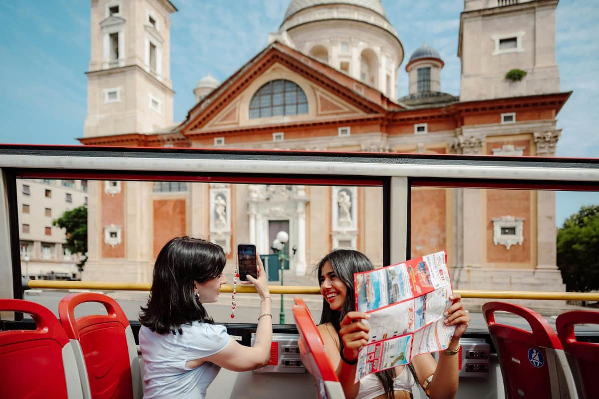Deux femmes dans un bus touristique, l'une tenant une carte et souriant, l'autre prenant une photo, avec un bâtiment historique en arrière-plan.