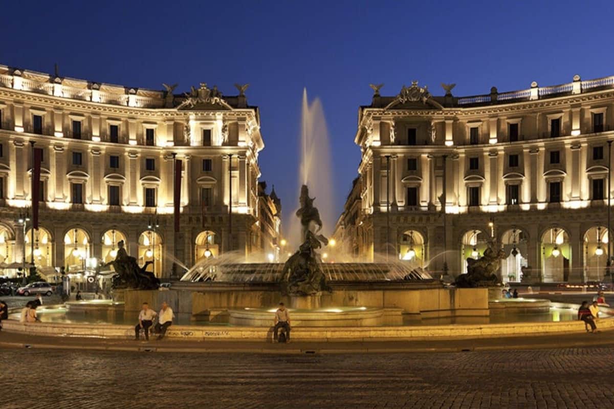 Des bâtiments illuminés et une fontaine avec des statues la nuit, avec quelques personnes assises à proximité sur une place publique.