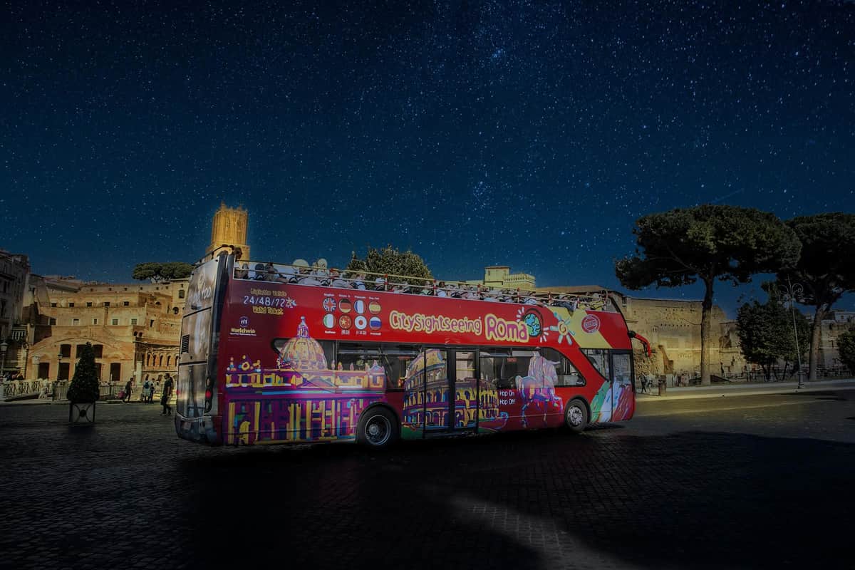 Rome : Visite nocturne en bus Bus touristique rouge à impériale à Rome, de nuit, avec un ciel étoilé et des bâtiments historiques en arrière-plan.