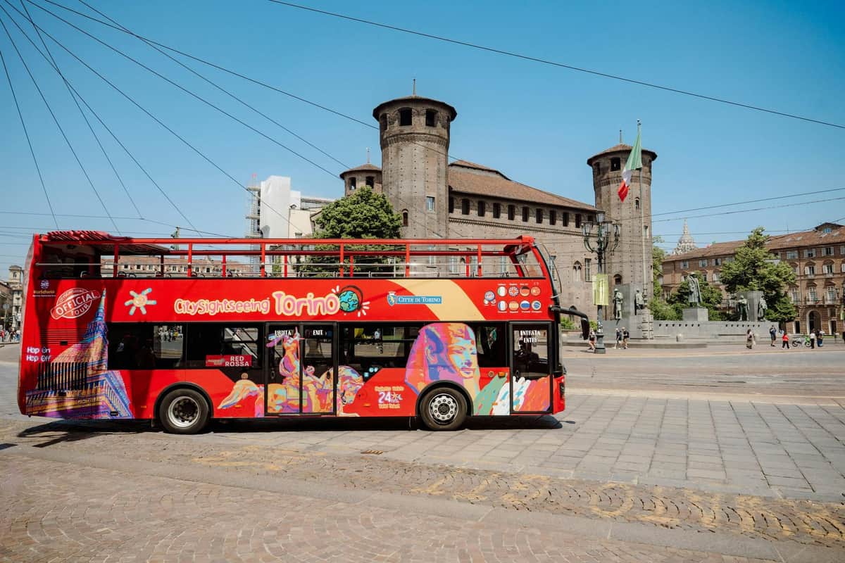 Un bus touristique rouge à impériale devant un vieux château en briques avec des tours rondes dans un paysage urbain ensoleillé.