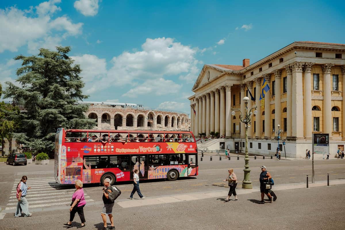 Un bus touristique rouge à impériale devant un bâtiment néoclassique et des ruines anciennes, par une journée ensoleillée.