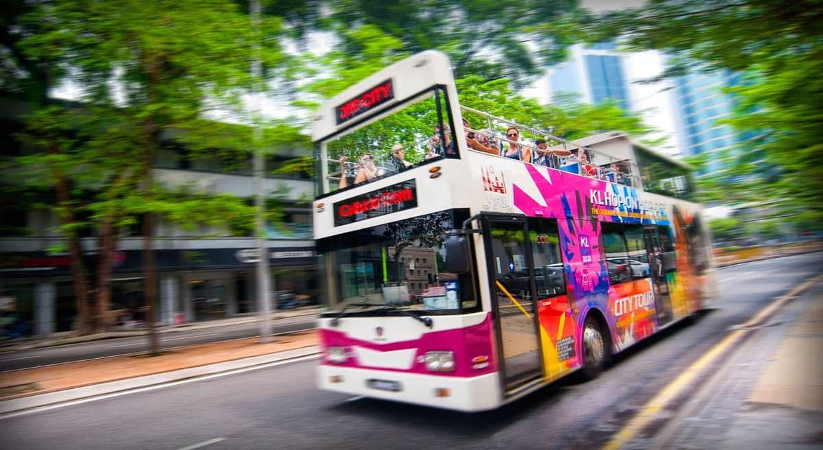 Un bus à impériale avec des publicités vibrantes et des touristes sur le pont supérieur circule dans une rue de la ville bordée d'arbres.