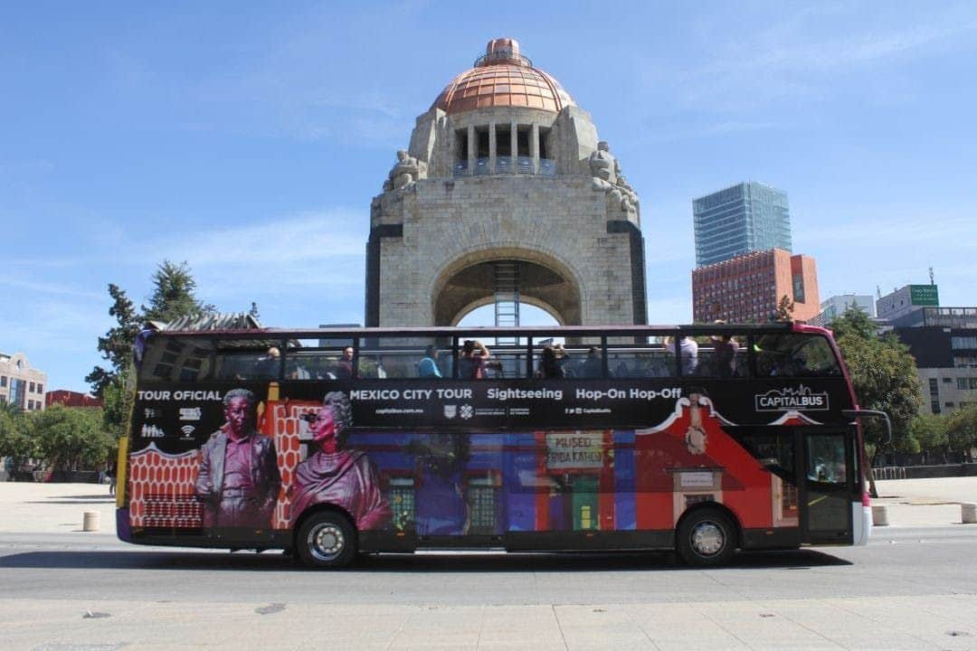Un bus touristique à impériale devant un monument historique sous un ciel bleu clair, avec des personnes sur le pont supérieur du bus.