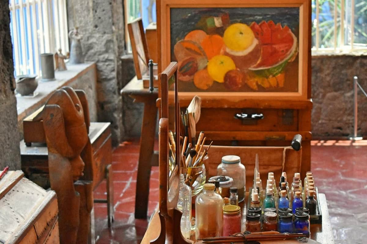 Un atelier d'artiste avec des bouteilles de peinture, des pinceaux et une palette. Une peinture de fruits colorée est exposée sur un chevalet à l'arrière-plan.