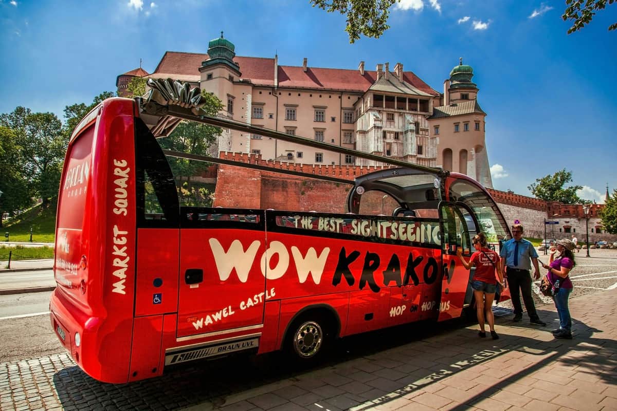 Bus touristique rouge devant un château historique sous un ciel bleu ; deux personnes debout à côté du bus.