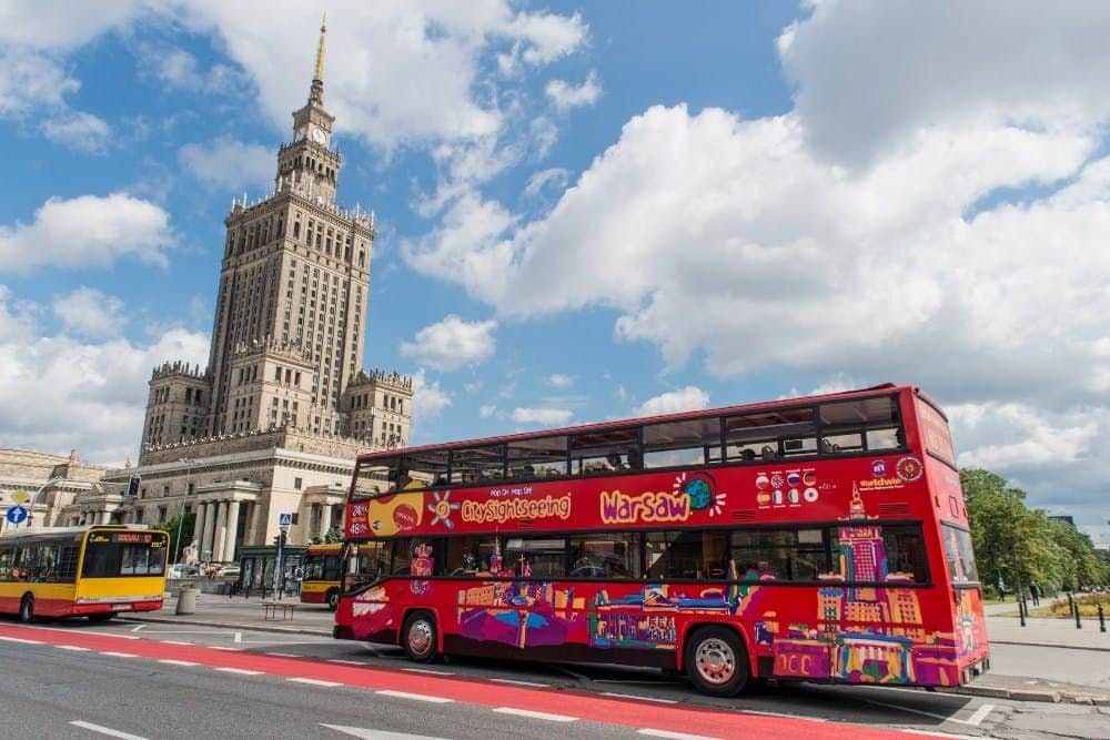 Bus hop-on hop-off Varsovie Bus touristique rouge à deux étages à Varsovie, avec un grand bâtiment historique en arrière-plan, sous un ciel partiellement nuageux.