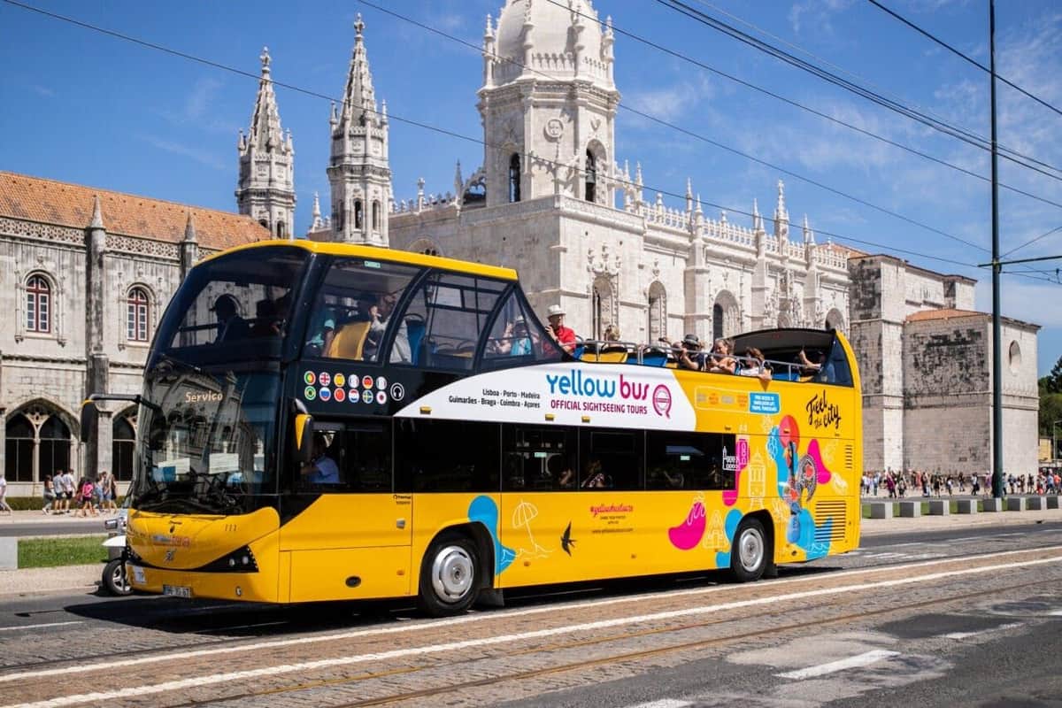 Ein gelber Doppeldecker-Sightseeing-Bus mit Fahrgästen vor einem historischen weißen Steingebäude unter einem klaren Himmel.