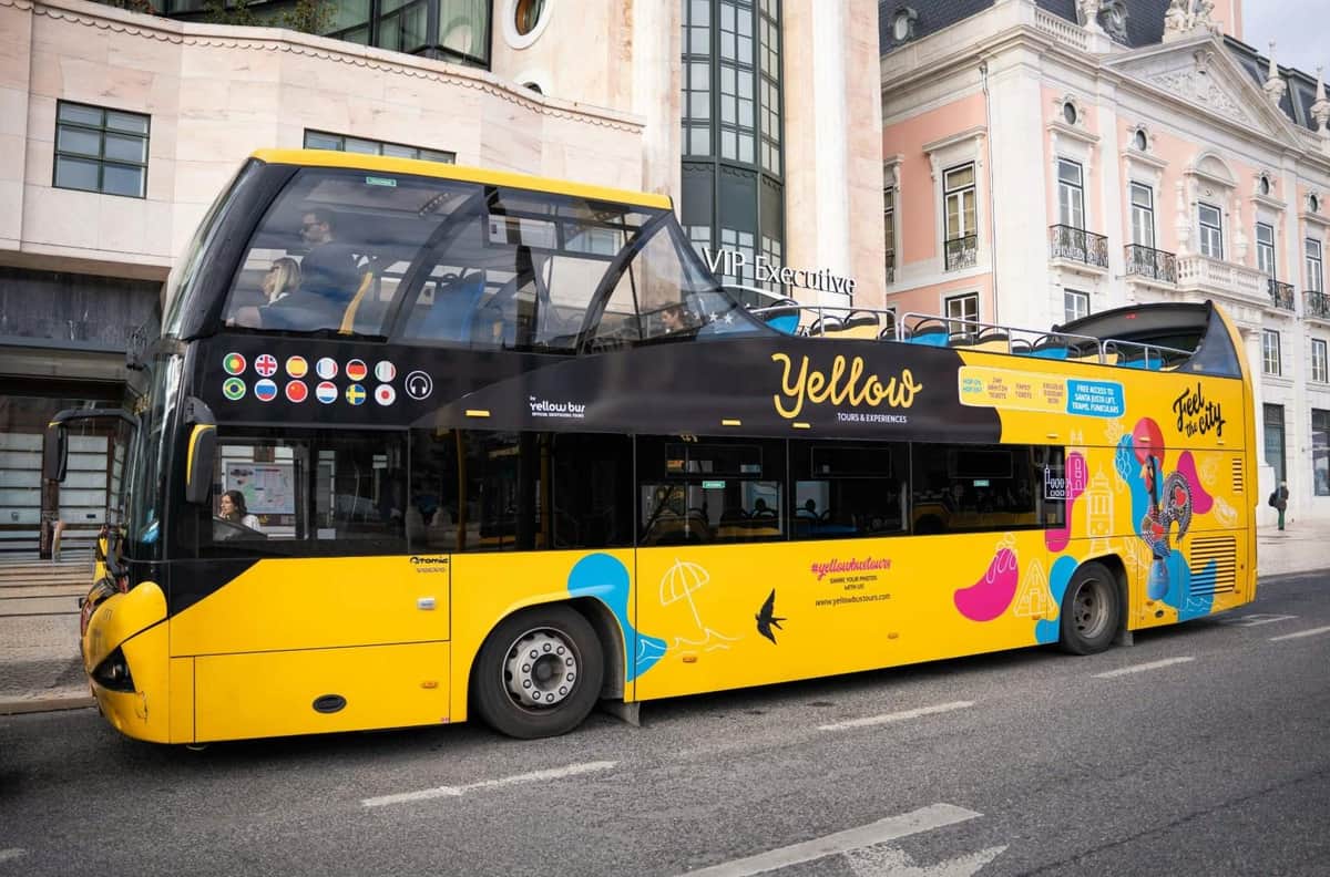 Un bus touristique à impériale jaune vif et noir, orné de motifs promotionnels, stationné devant un bâtiment dans une rue de la ville.