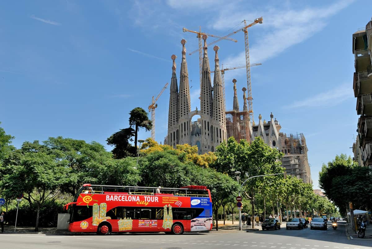 Un bus touristique rouge à impériale devant la Sagrada Familia, avec des grues et des arbres sous un ciel bleu.