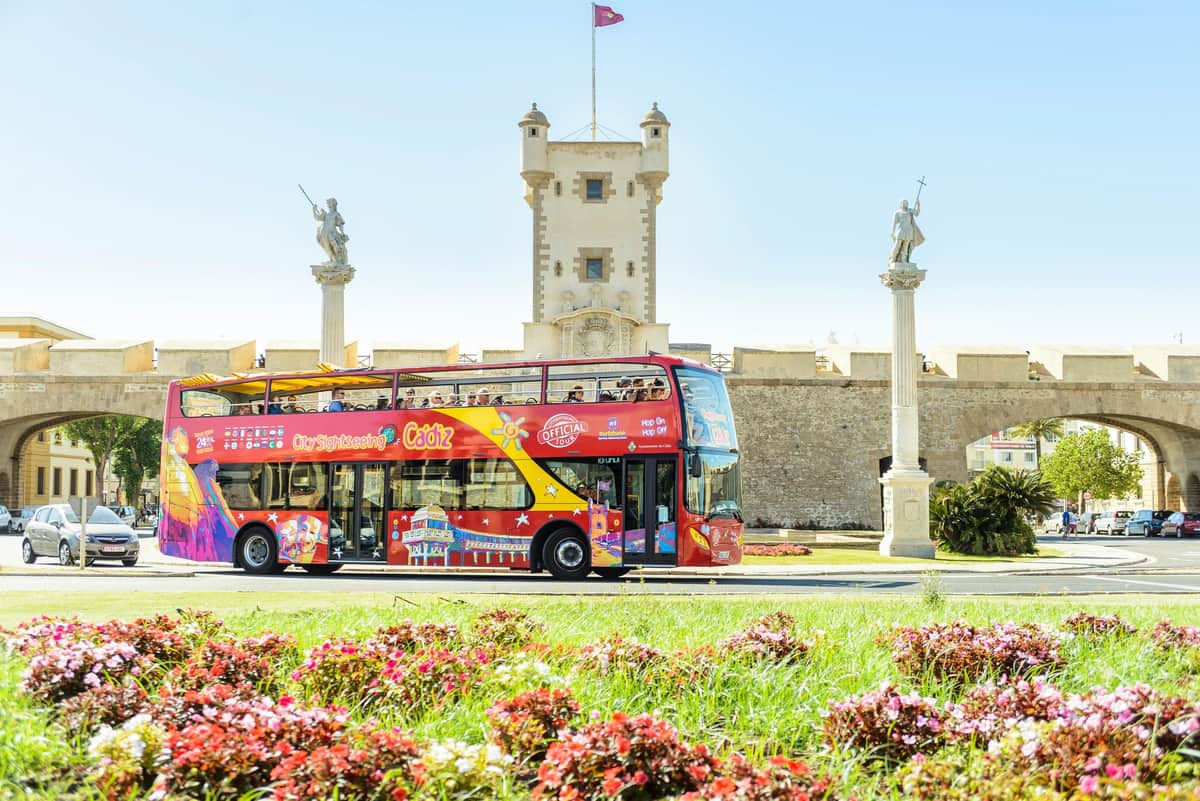 Bus hop-on hop-off Cadix Un bus touristique rouge à impériale passe devant un bâtiment historique orné de statues et d'un drapeau, entouré de fleurs et de verdure éclatantes.