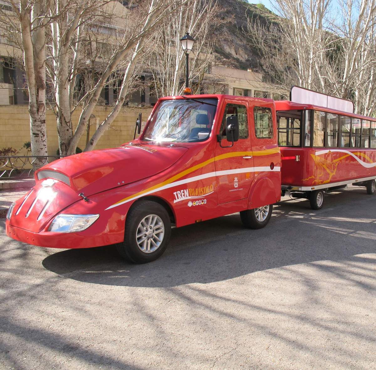 Un train touristique rouge vif avec une cabine et des wagons de passagers ouverts sur une rue bordée d'arbres sans feuilles.