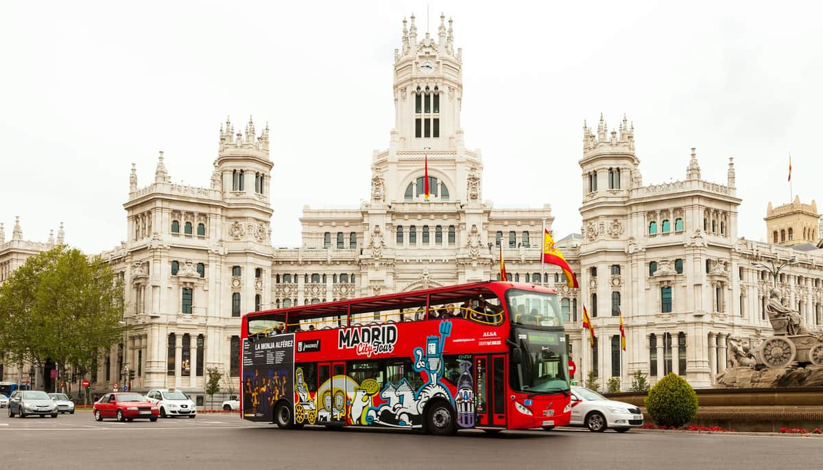 Un bus rouge à impériale portant l'inscription "Visite en espagnol" devant un bâtiment blanc orné de drapeaux espagnols.