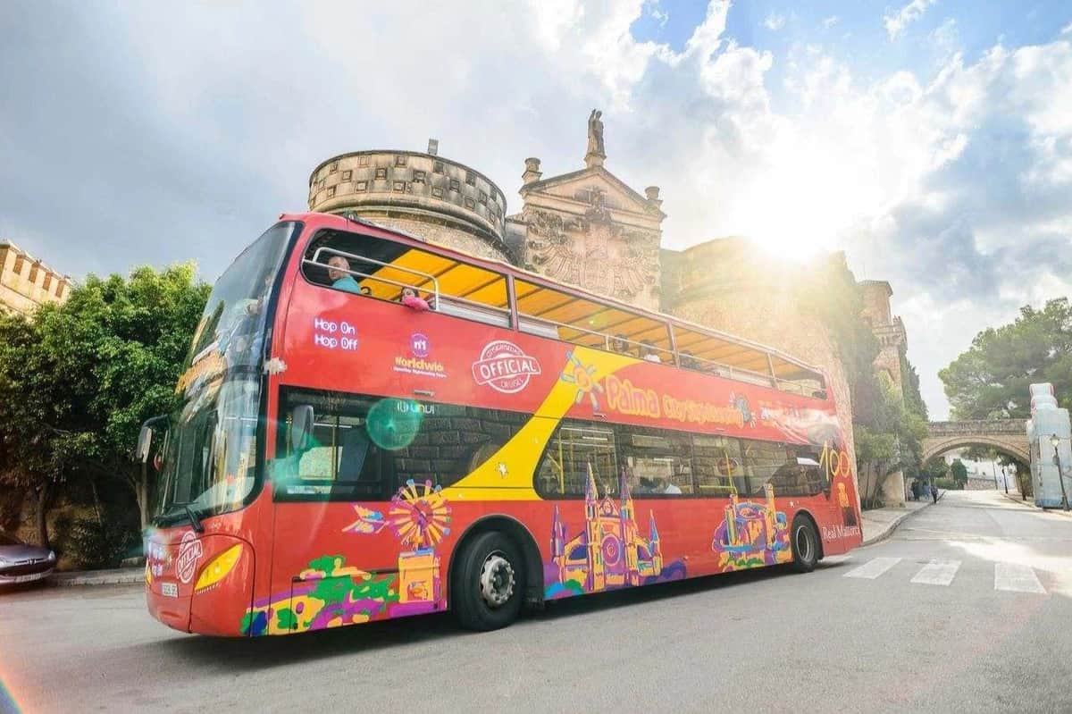 Un bus touristique rouge à impériale avec des graphiques colorés sur le côté, garé près de bâtiments historiques sous un ciel partiellement nuageux.