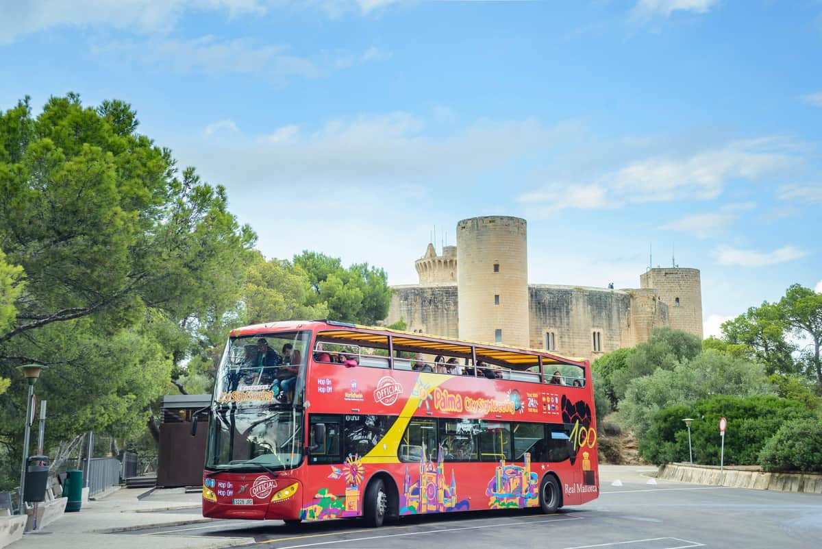 Un bus touristique rouge à impériale, aux graphismes éclatants, est garé près d'un château en pierre entouré d'arbres, sous un ciel partiellement nuageux.