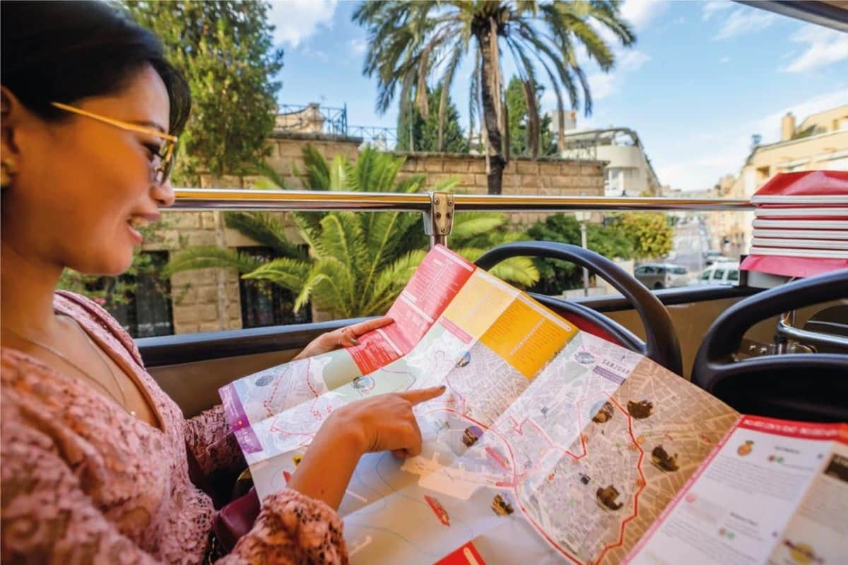 Une femme tenant et montrant une carte, assise dans un bus à toit ouvert avec des palmiers et des bâtiments historiques en arrière-plan.