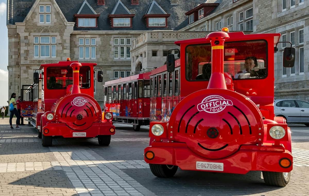 Santander : Bus-on Hop-off + Train Tour Deux trains touristiques rouges portant le logo "Visite officielle" sont garés à l'extérieur d'un grand bâtiment en pierre orné de multiples fenêtres et pignons.