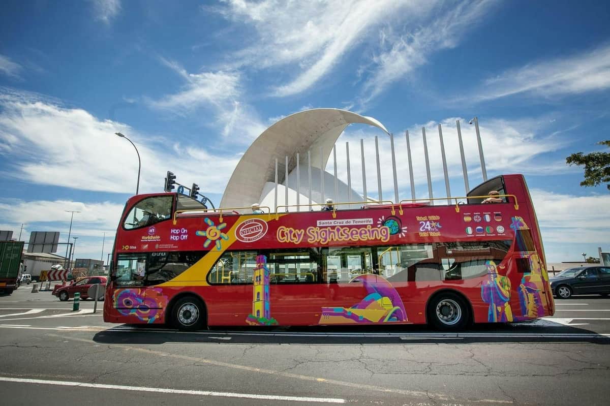 Santa Cruz de Tenerife : Circuit en bus Hop-on Hop-off de 24 ou 48 heures Un bus touristique urbain rouge à impériale se tient devant une structure architecturale moderne et blanche, sous un ciel partiellement nuageux.