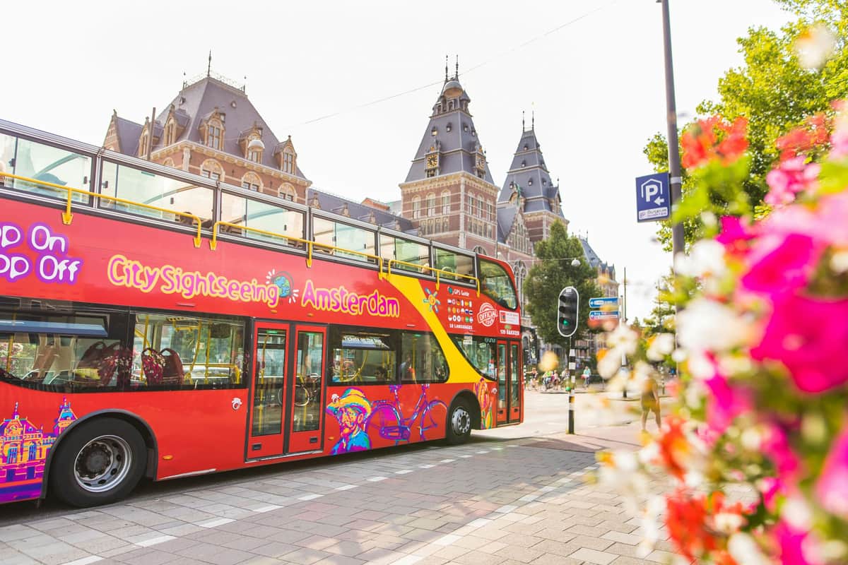 Un bus touristique rouge à impériale portant l'inscription "City Sightseeing Amsterdam" est garé près de bâtiments historiques lors d'une journée ensoleillée.