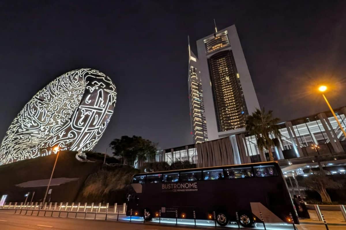 Bustronome Dubaï : Visite en bus + dîner Vue nocturne d'un bâtiment moderne, d'une sculpture illuminée aux motifs complexes et d'un bus portant l'inscription "BUSTRONOME".