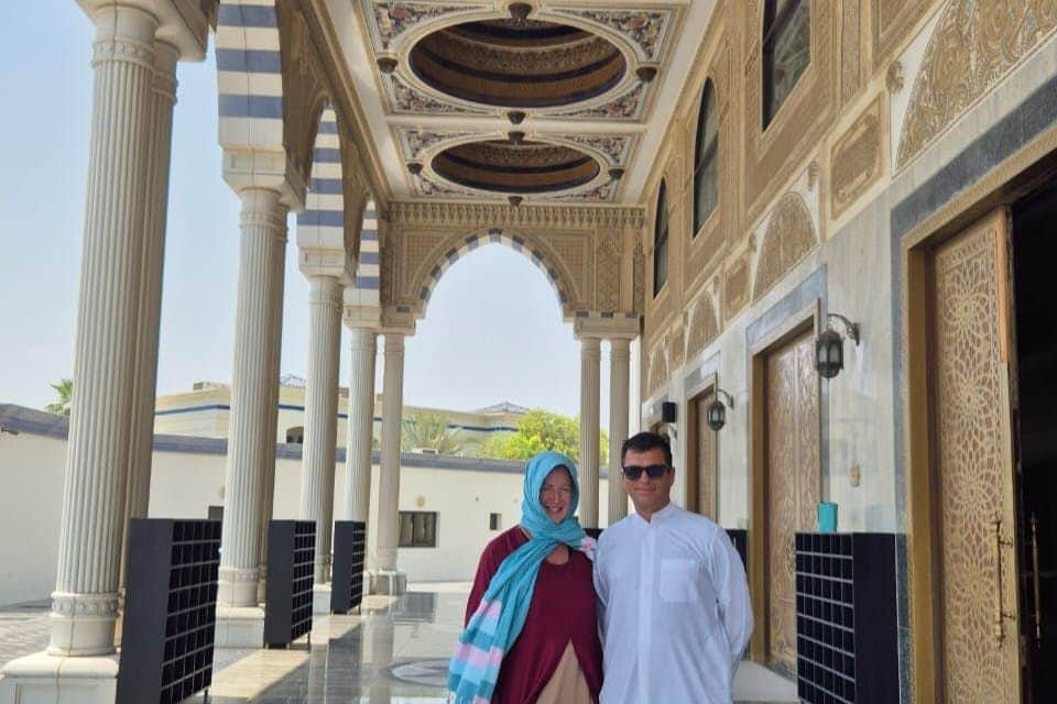 Deux personnes se tiennent dans un grand couloir architectural avec des plafonds ornés, des colonnes et des portes cintrées. L'une porte des lunettes de soleil, l'autre un foulard.