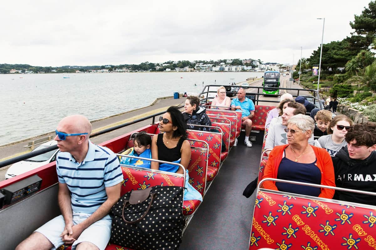 Personnes assises sur le pont supérieur d'un bus touristique aux sièges à motifs rouges, voyageant le long d'une route côtière où l'on aperçoit l'eau et les bâtiments.