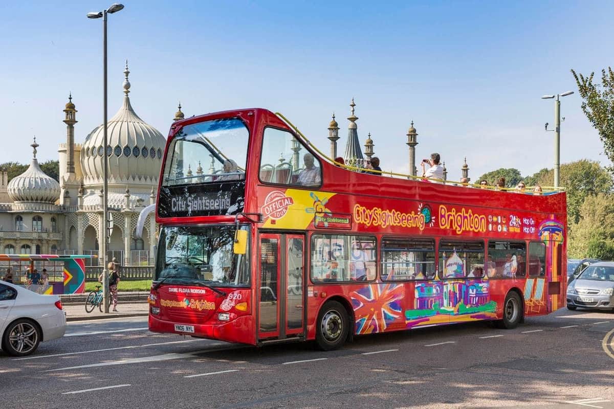 Bus Hop-on Hop-off Brighton Bus touristique rouge à impériale à Brighton avec des touristes sur le pont supérieur, passant devant un bâtiment à l'architecture en dôme.