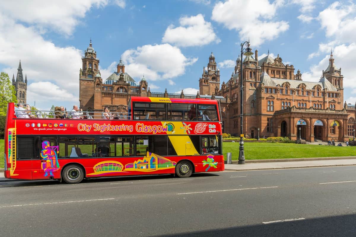 Bus hop-on hop-off Glasgow Un bus touristique rouge à impériale portant l'inscription "City Sightseeing Glasgow" passe devant un bâtiment historique sous un ciel partiellement nuageux.